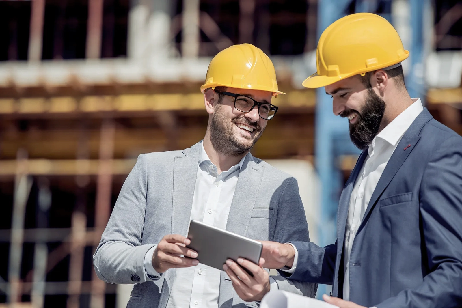 Two men wearing blazers and yellow hard hats talk as they look at a tablet on a construction site.