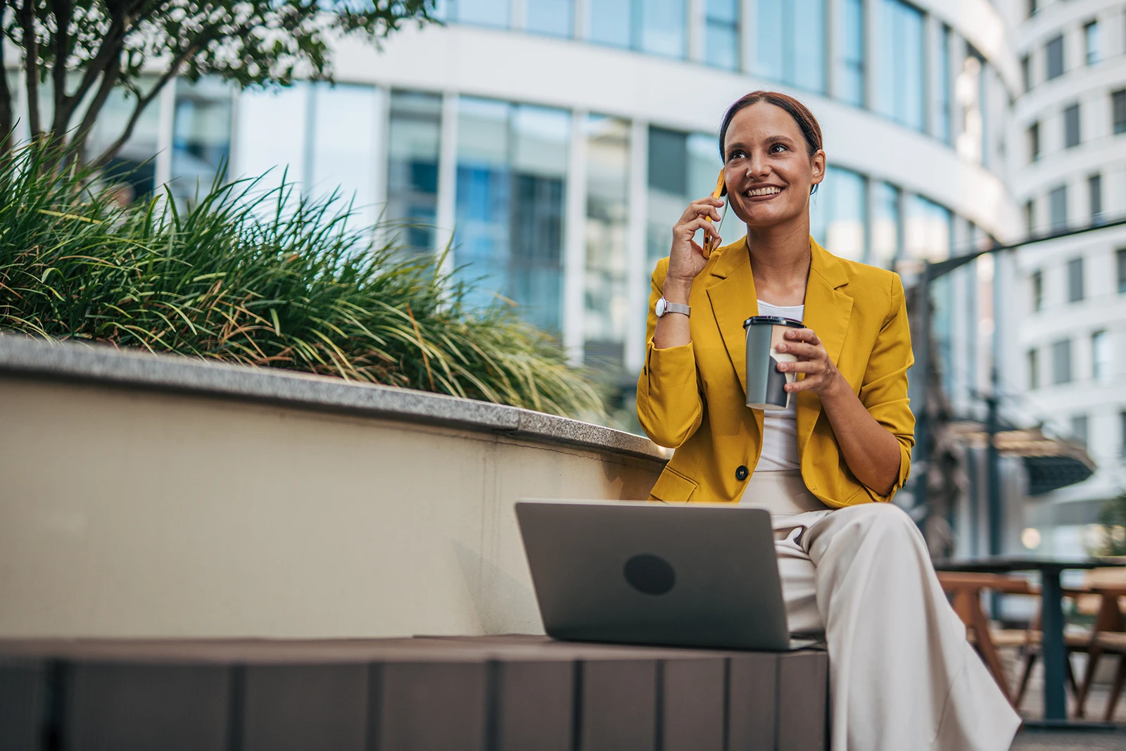 A woman in a yellow blazer sits outside on a patio with her laptop open in front of her. She has a coffee in hand and talks on her cell phone.