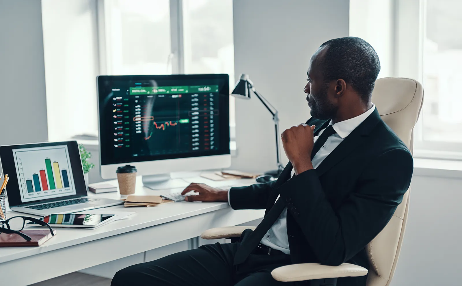 Dans un bureau, un homme en complet est assis à son bureau, devant un ordinateur portable et un grand écran. Il regarde les graphiques boursiers sur l'écran.