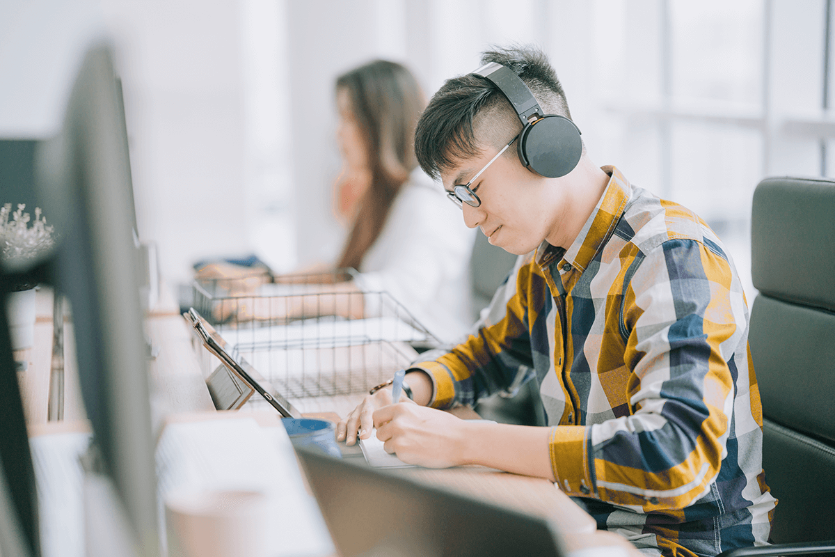 A student sits at a shared desk in front of a computer monitor, wearing headphones and writing in a notebook. Another student studies in the background.