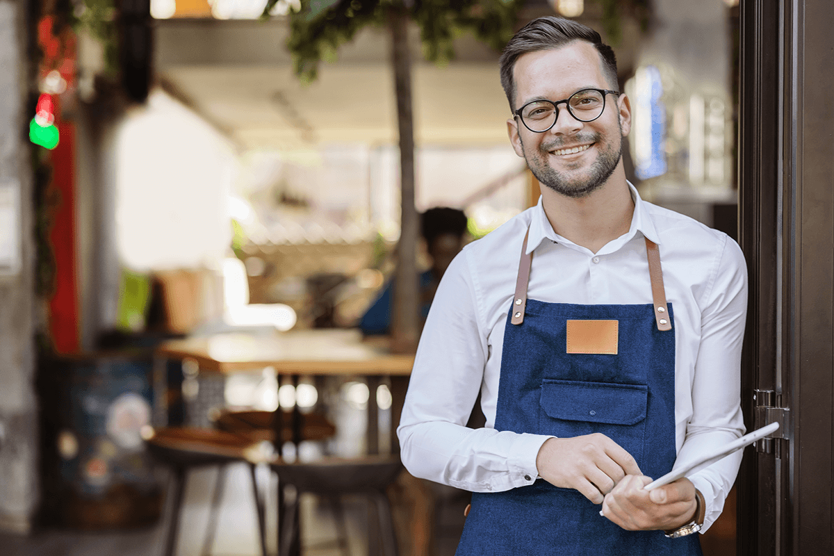 A young man wearing an apron smiles as he looks up from his tablet and out his restaurant door.
