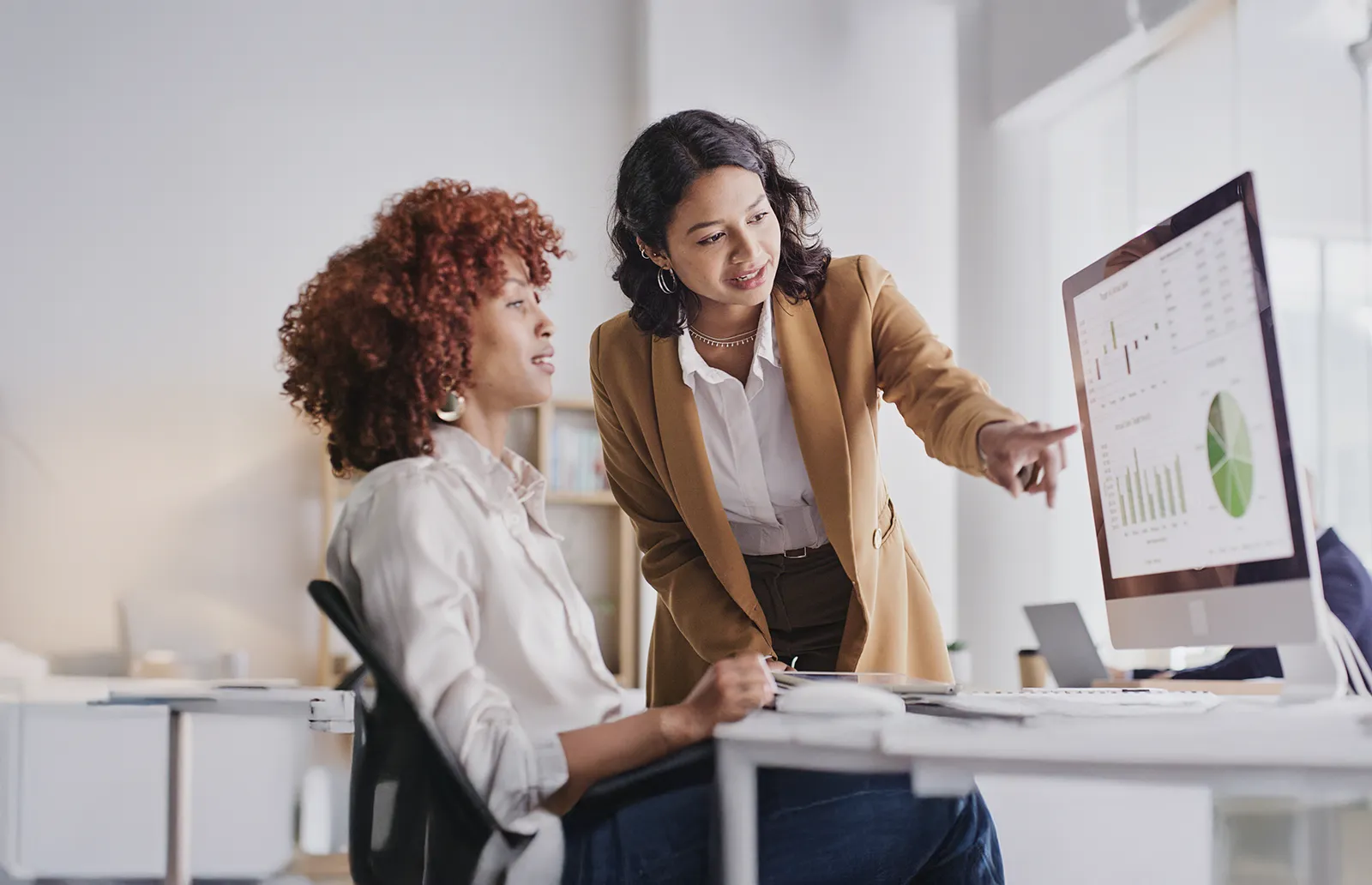 Deux femmes dans un bureau discutent devant un ordinateur qui affichent des graphiques financiers.