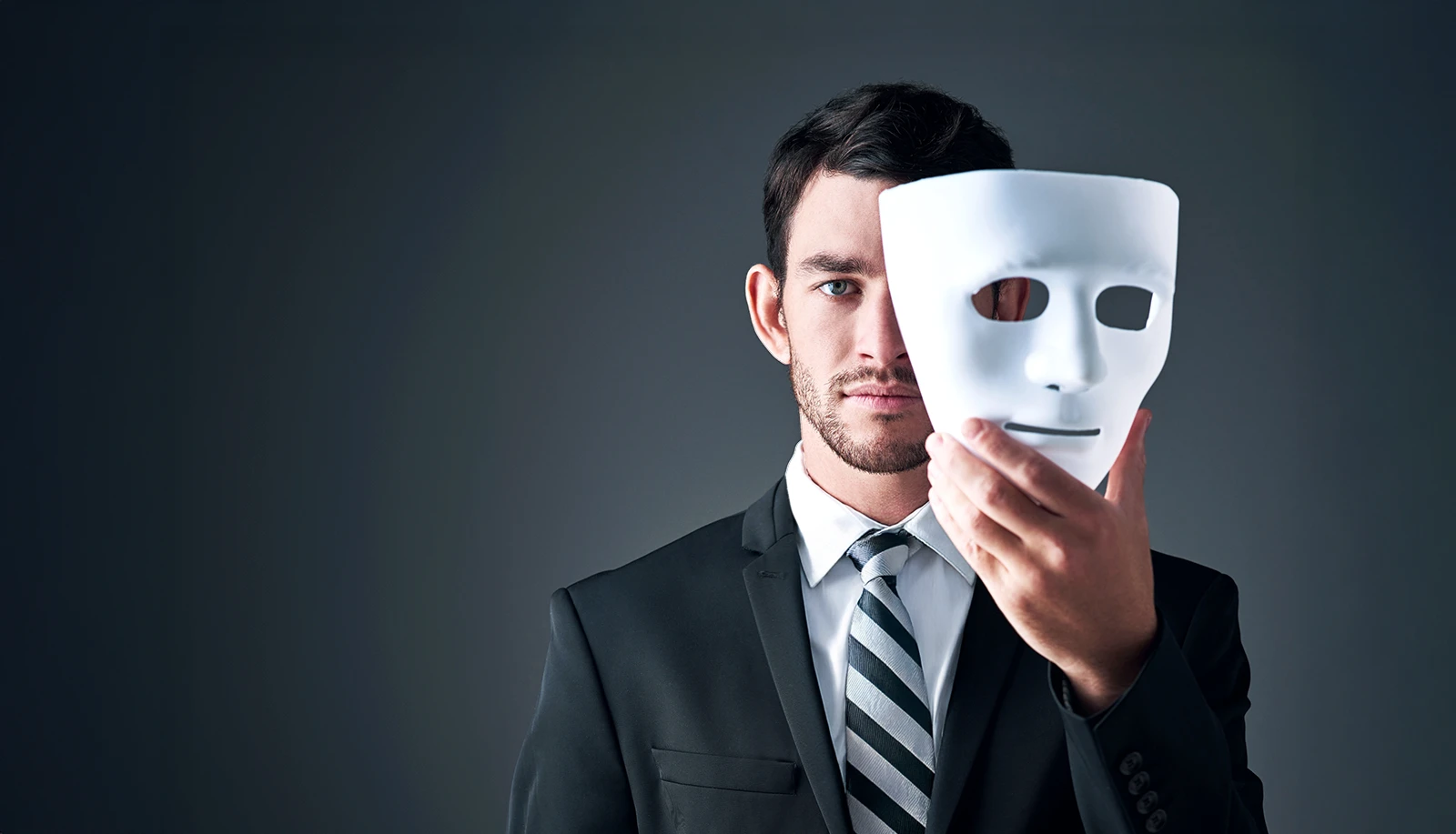 A man wearing a suit and tie holds a white mask covering half of his face.