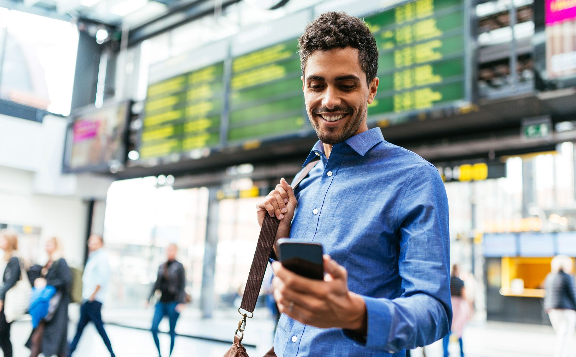 A man stands in a train station with a duffle bag on his should while smiling down at his cell phone