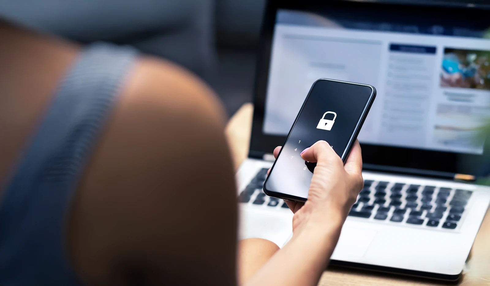 Gros plan d'une personne tenant un téléphone intelligent verrouillé avec un ordinateur portable ouvert en arrière-plan.