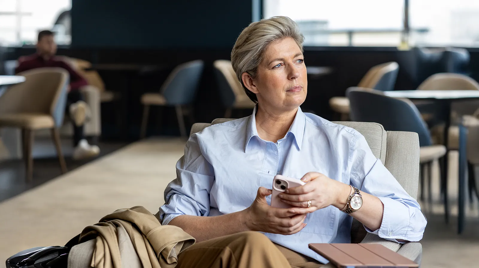 Une femme d'affaires assise sur un fauteuil tient son téléphone mobile des deux mains et regarde au loin.