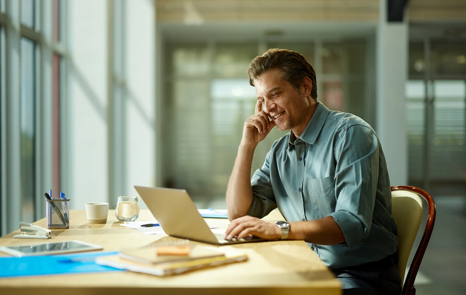 A man sits at a desk in front of an open laptop, smiling as he looks at the screen.