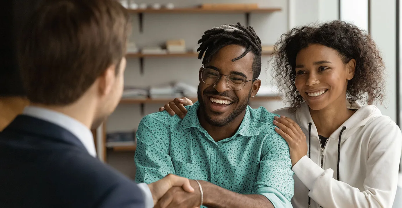 A young smiling couple sit at a wooden desk with paperwork in front of them. The man holds a pen and is shaking hands with another man in a suit.