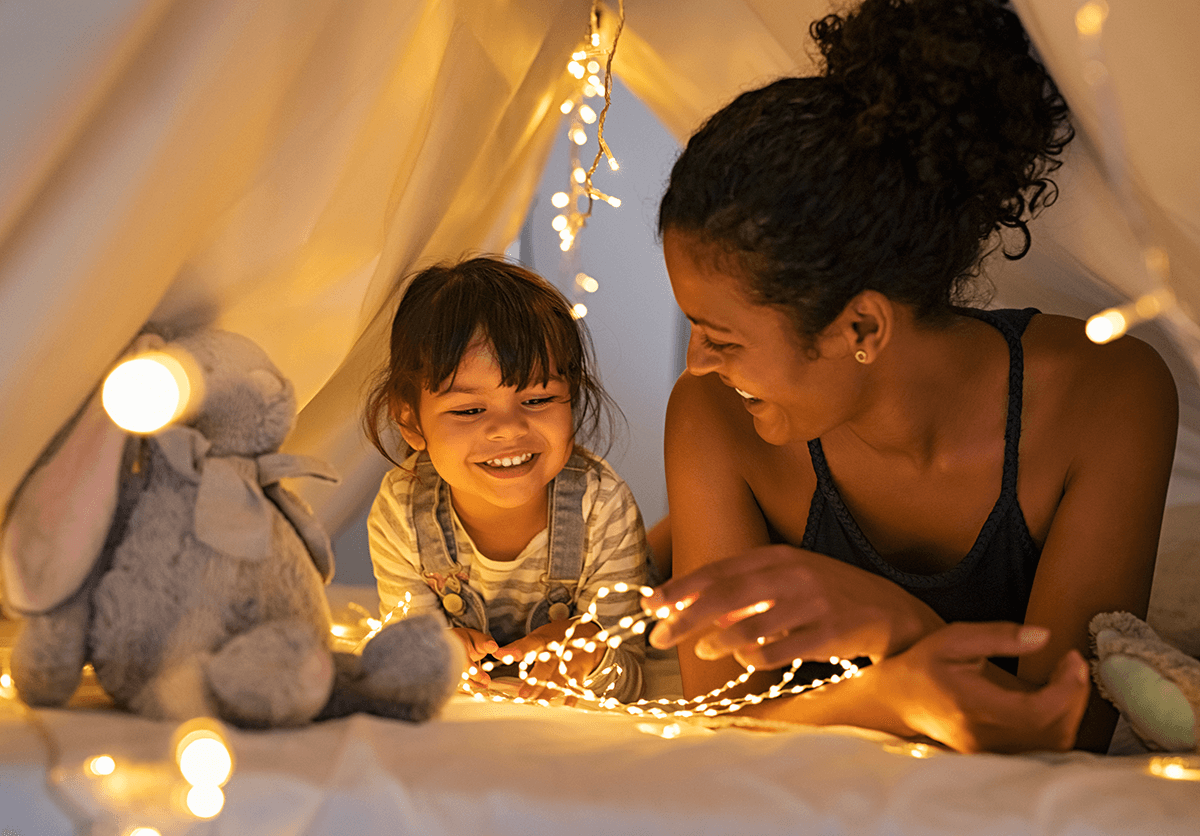 A mother and daughter are in a cloth tent set up indoors. Surrounded by soft fairy lights, they lay happily on their stomachs, propped up on their arms.