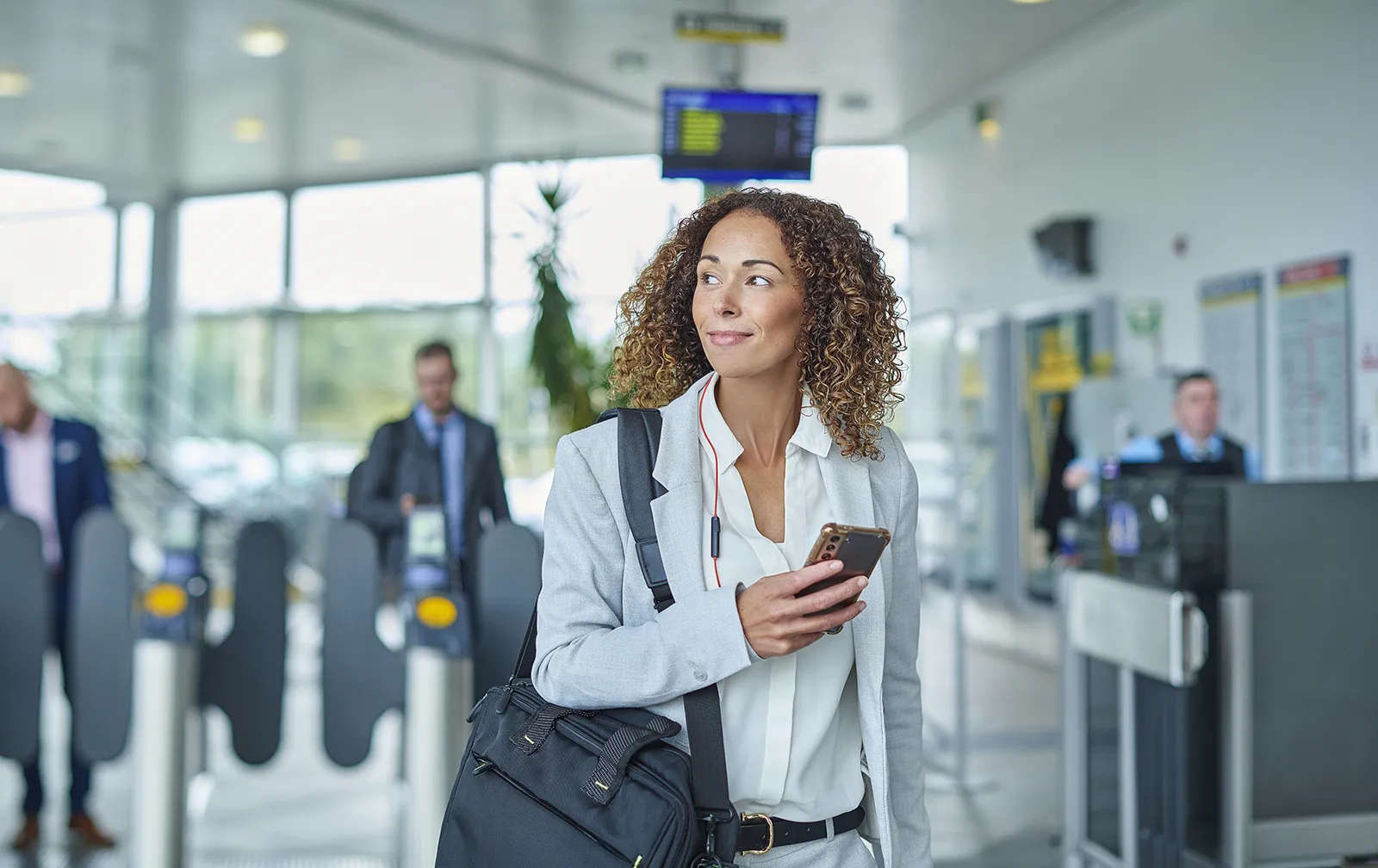 Une femme en tenue d’affaires est dans une gare. Elle tient son téléphone portable en main et elle porte en bandoulière un sac d’ordinateur.