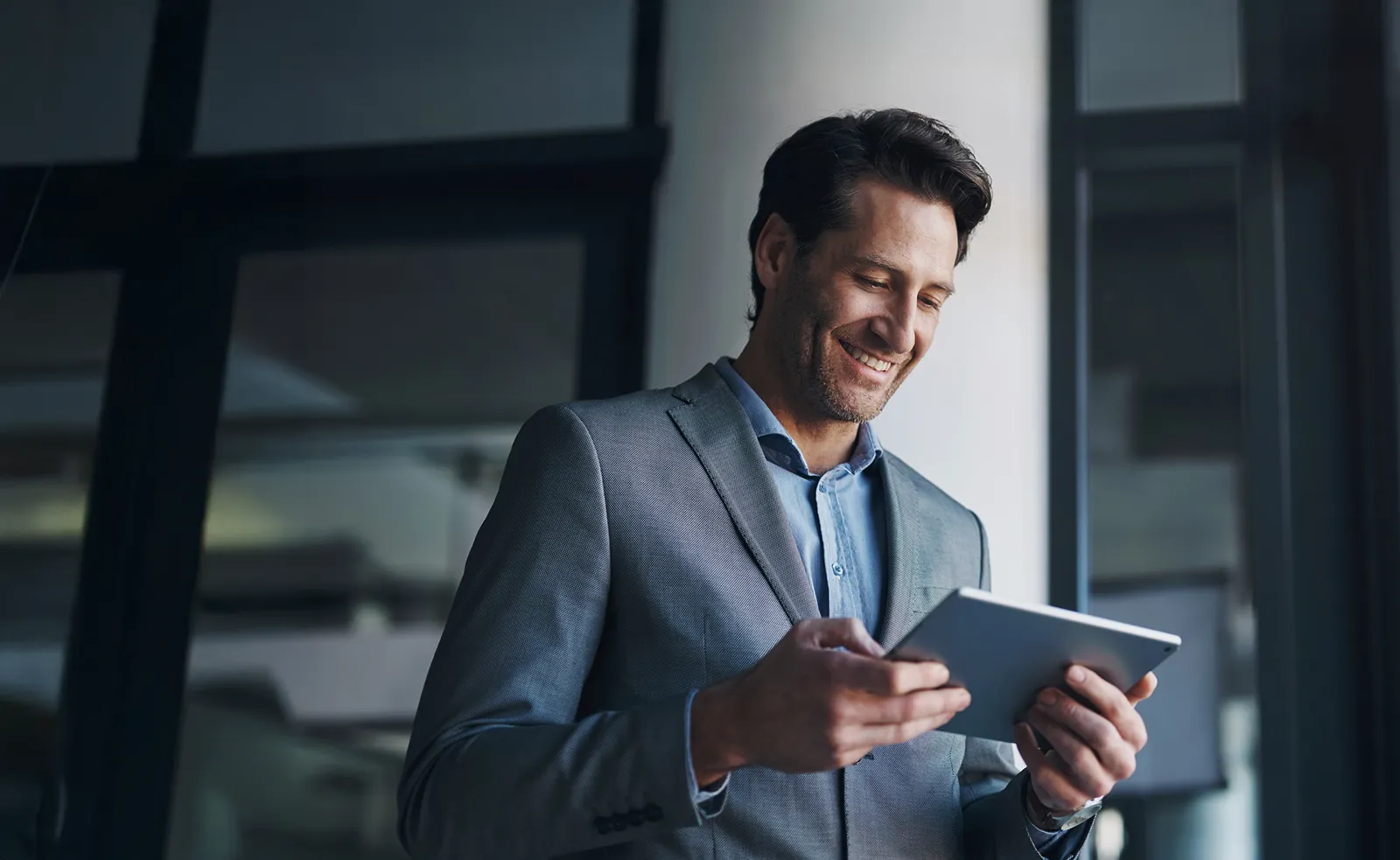 A man dressed in a suit jacket smiles while looking at a tablet in his hands.