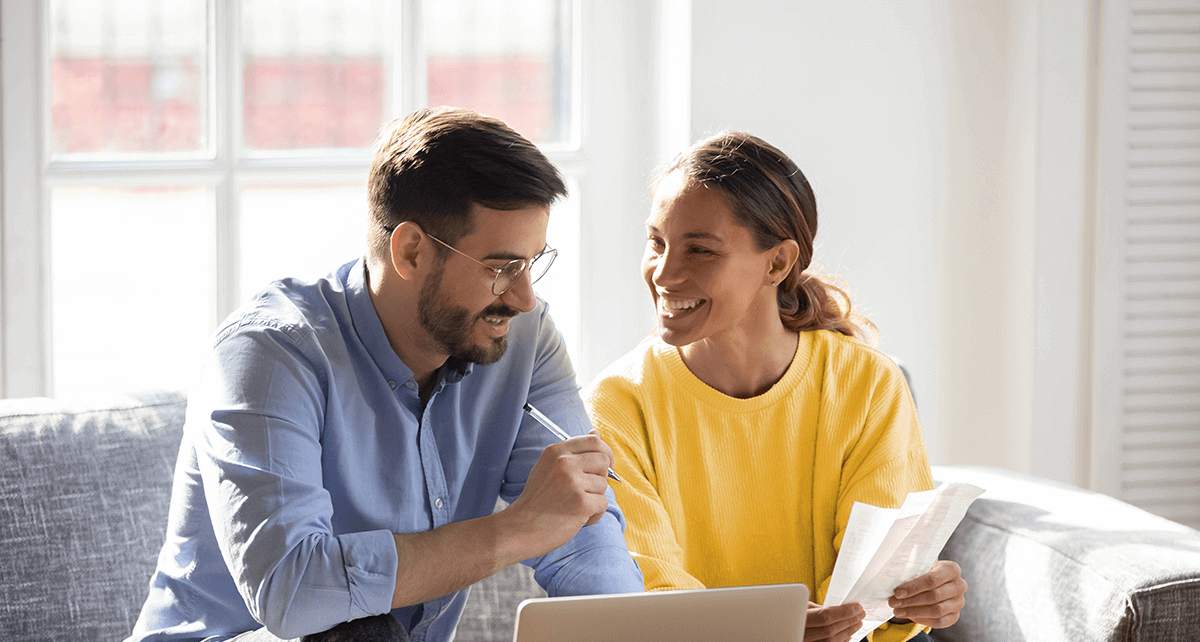 Un couple est assis sur un canapé, avec un ordinateur portable, des documents et une calculatrice posés sur la table devant eux.