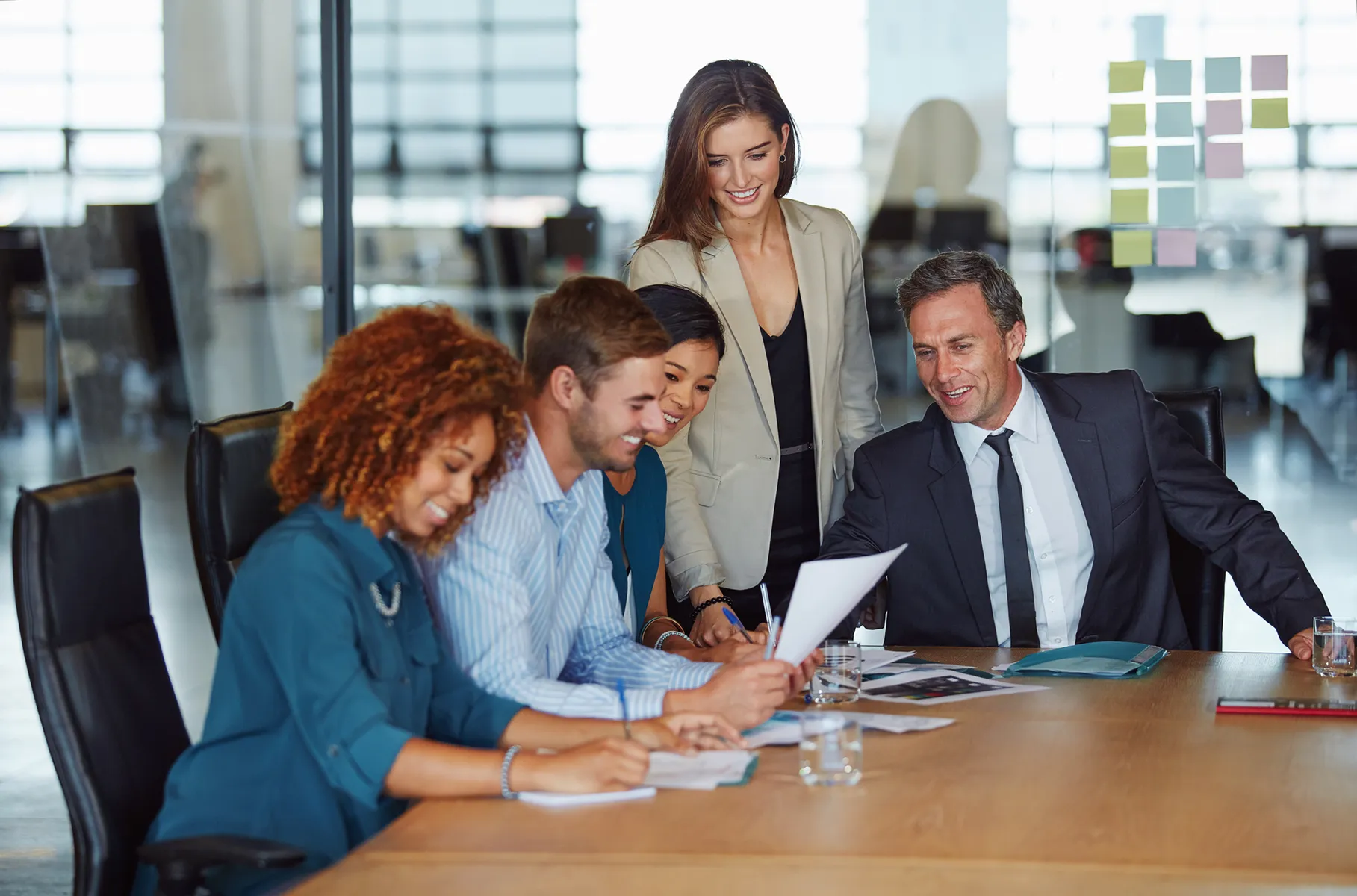 In a boardroom, a team of five people sit or stand around a big, wooden table.