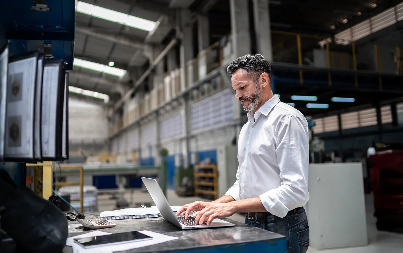 In a large warehouse, a man stands at a table, working on a laptop.