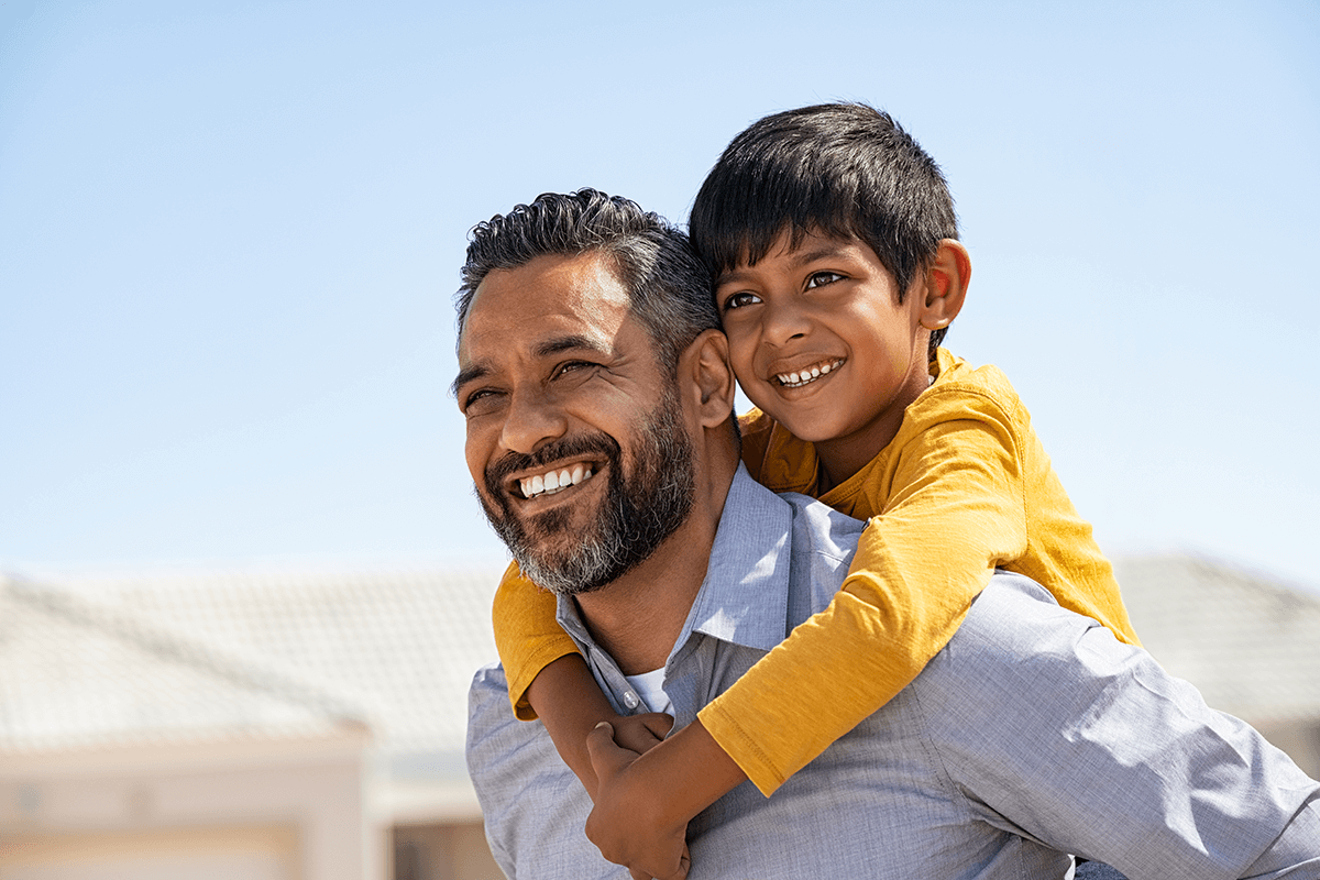 Père portant son fils sur le dos devant leur maison, profitant d'un moment de bonheur ensemble.