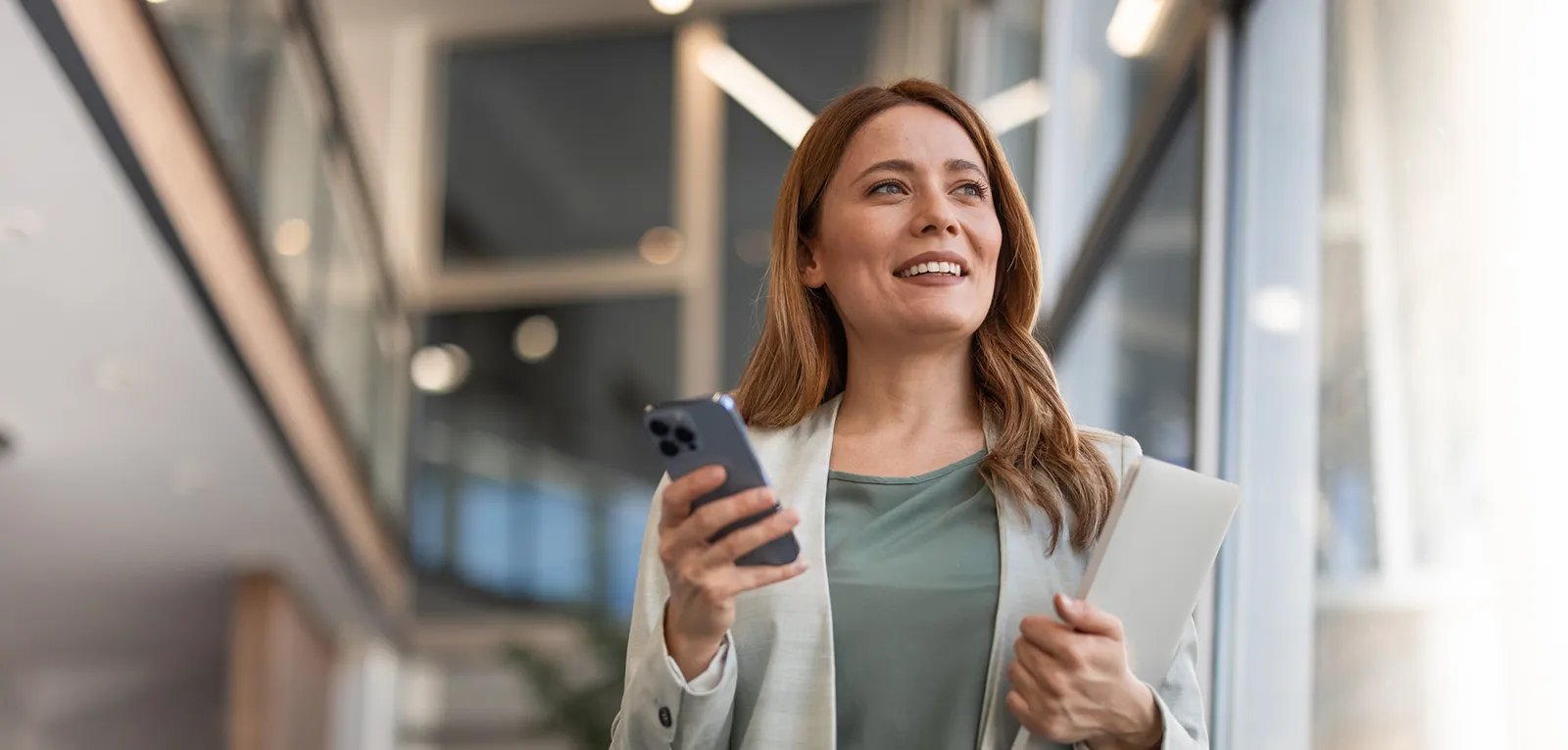 A businesswoman stands and smiles in a bright building lobby while holding a mobile phone and laptop.