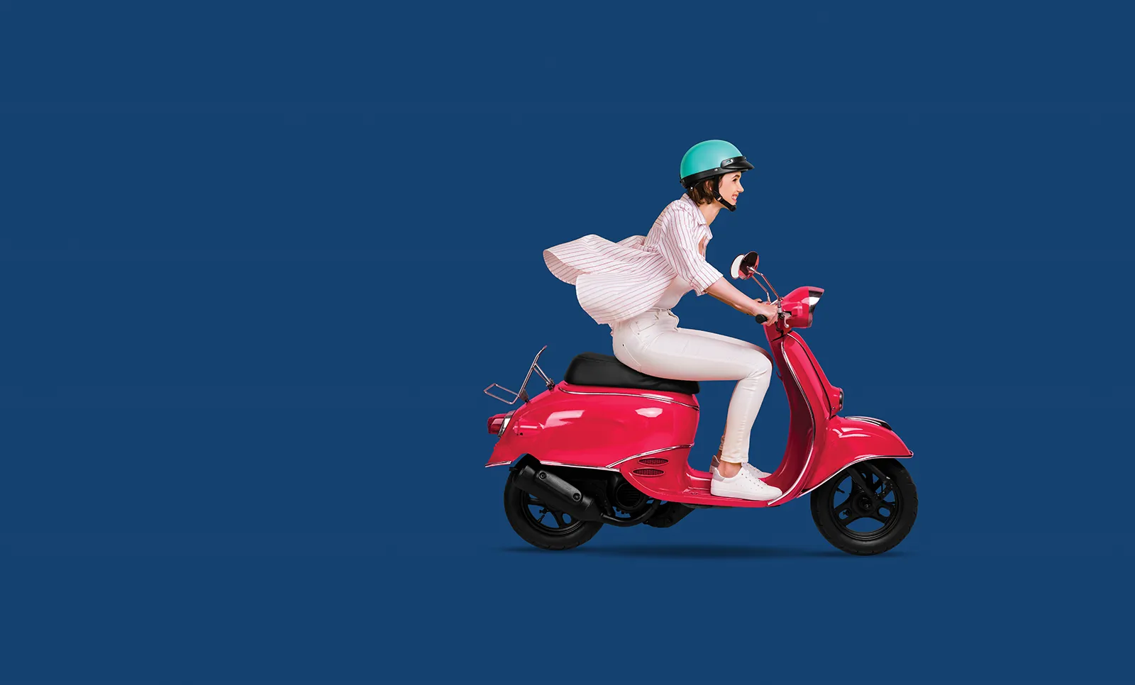 A woman wearing a turquoise helmet smiles as she rides a red scooter, her blouse billowing in the wind.