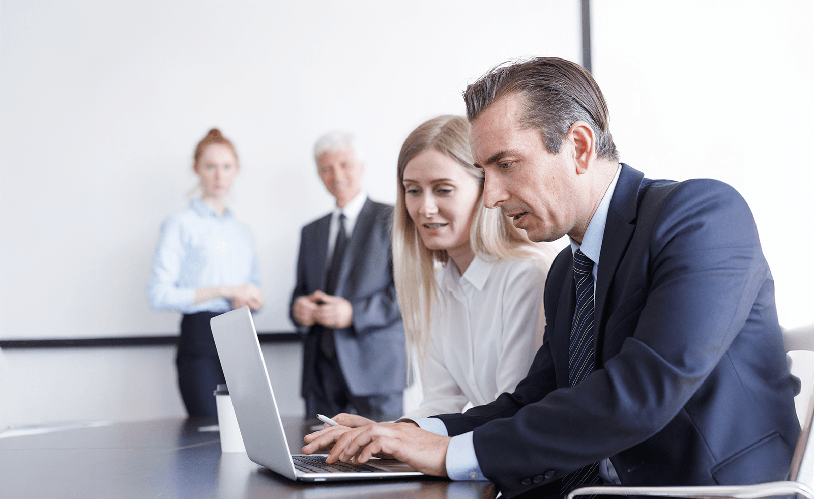 In a conference room, two professionals work at a laptop while two other people in the background look on.